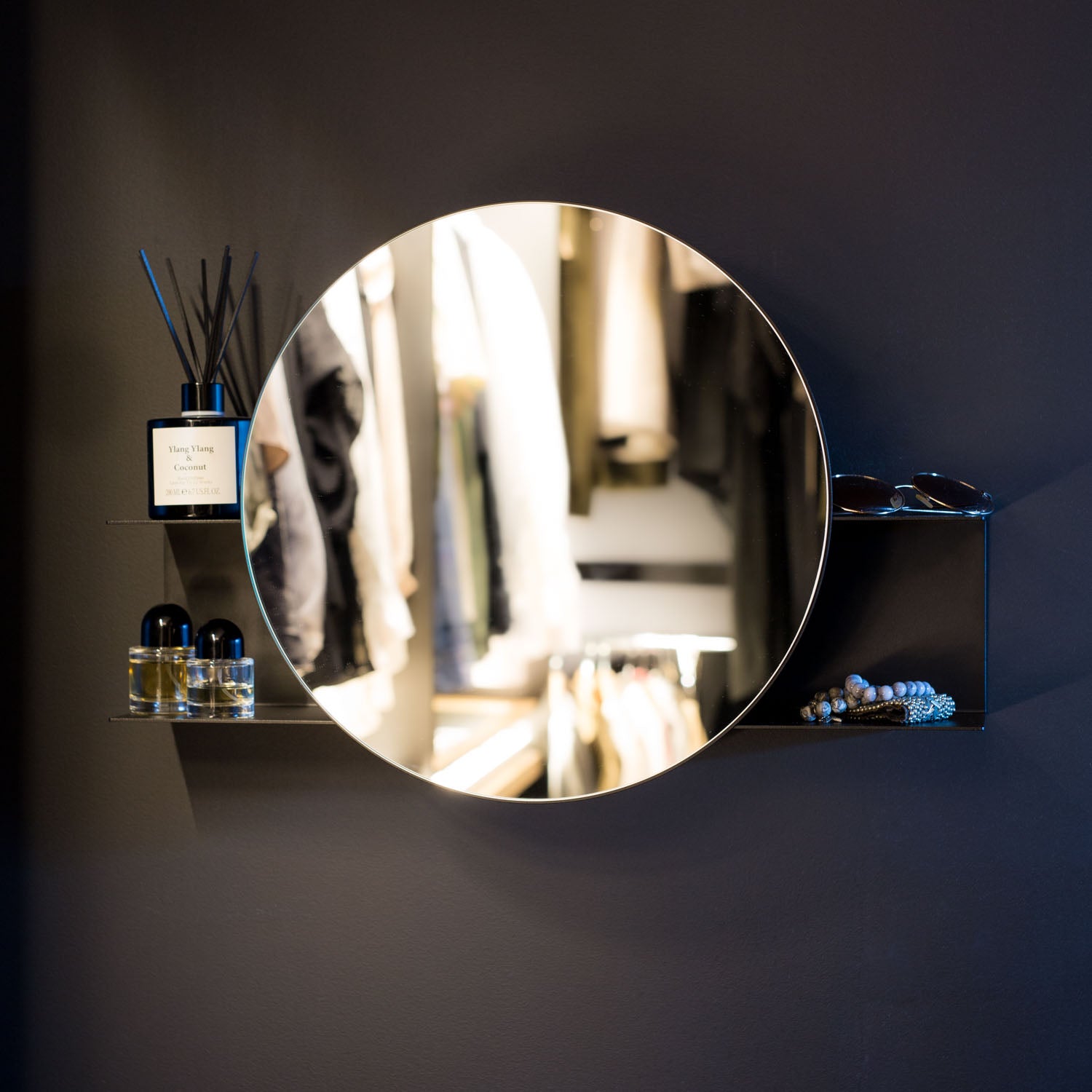 Round mirror with shelf on a dark wall with decorative items around it.